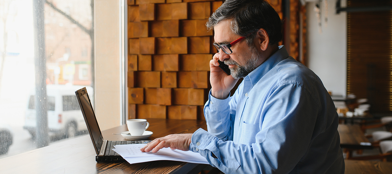 Senior man on cell phone, using laptop at a coffee shop